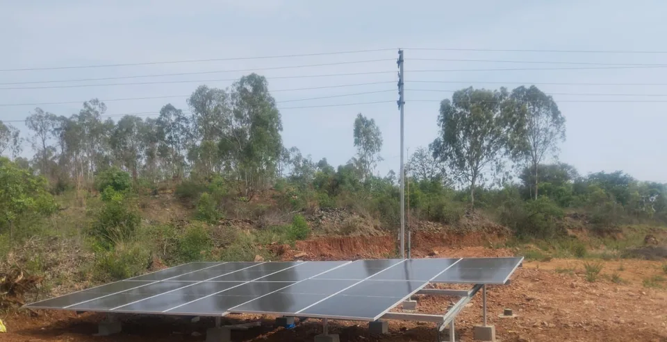 Ground-mounted solar array on red-earth terrain at Arakanakere, Karnataka, with eucalyptus trees and a power pole behind