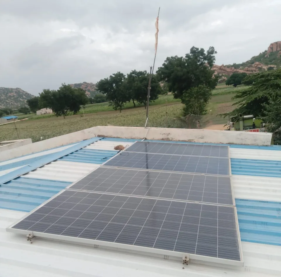 Solar panels on a corrugated metal rooftop at Nageshanahalli, Karnataka, with rocky hills and farmland beyond
