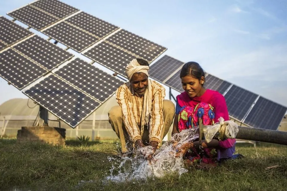 A man and a girl crouching at a borewell outlet as water pours out, a row of solar panels on a frame behind them.
