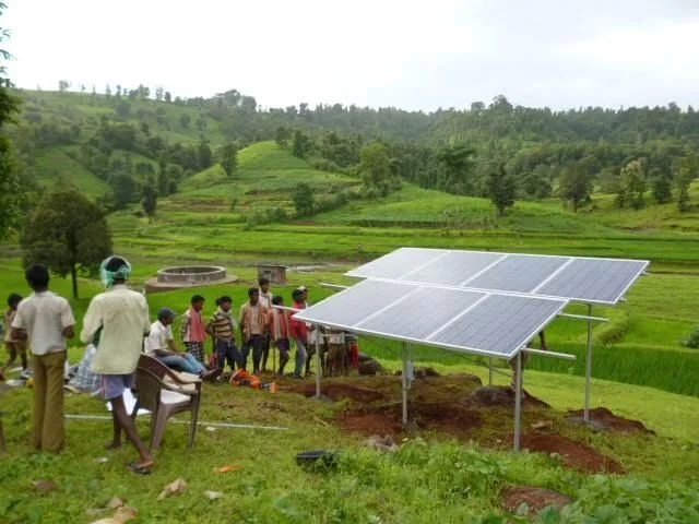 A ground-mounted solar array being installed on a green terraced hillside, with a group of people gathered around it.