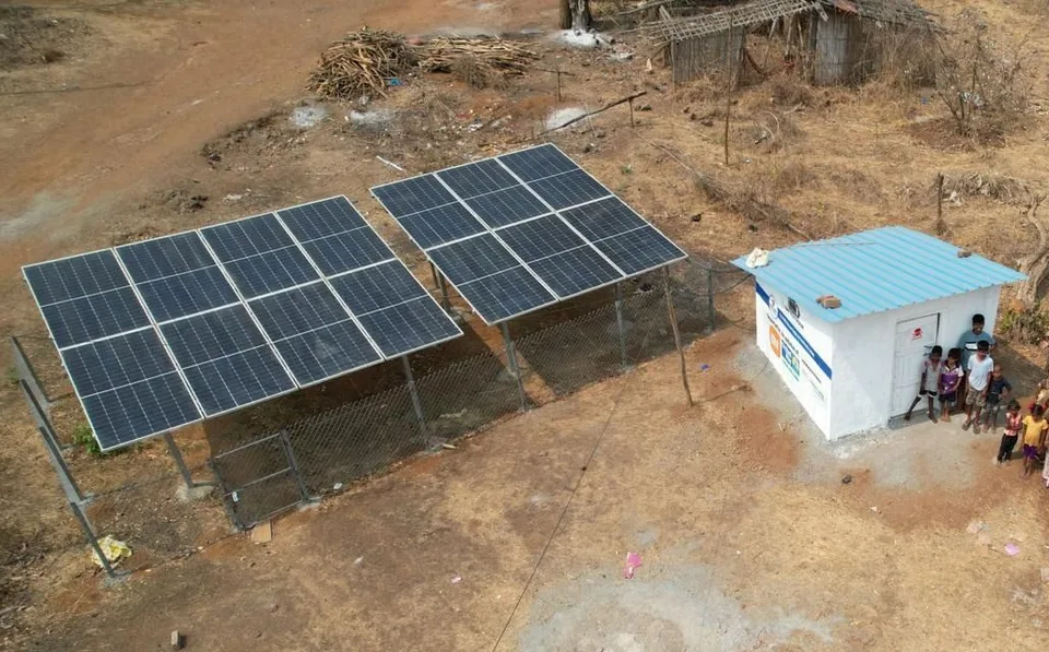 Two ground-mounted solar arrays on bare red earth beside a small white powerhouse with a blue metal roof, a group of villagers standing at its door.