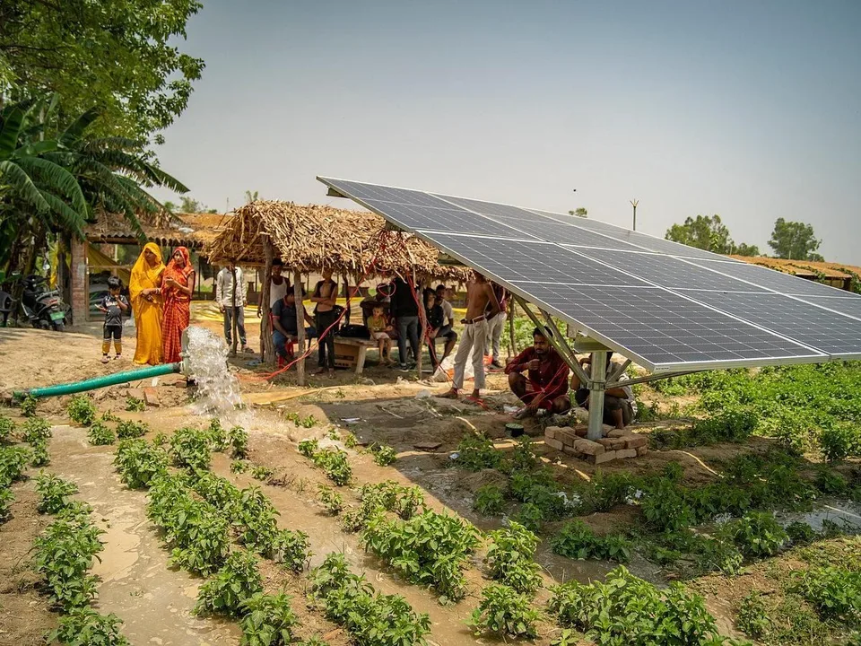 Water pouring from a pipe into a channel between rows of young crops, a solar panel on a raised frame beside it and people watching from a thatched shelter.