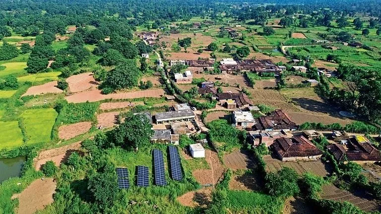 Aerial view of a village surrounded by fields and trees, with a small ground-mounted solar array beside the buildings.