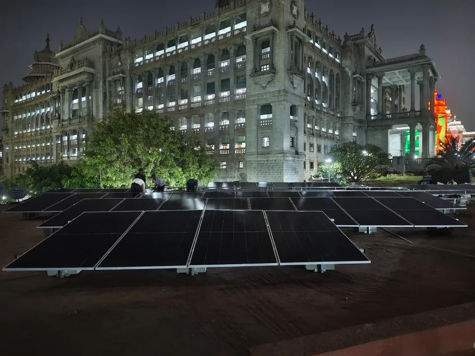 Modules being installed on a flat terrace after dark, with the floodlit granite facade of the Vikasa Soudha behind