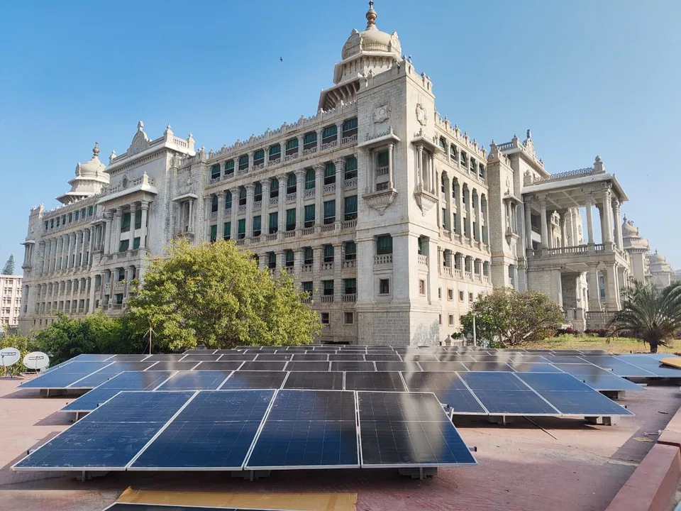 A long completed run of rooftop modules on the terrace, with the full stone facade of the Vikasa Soudha rising behind them in daylight