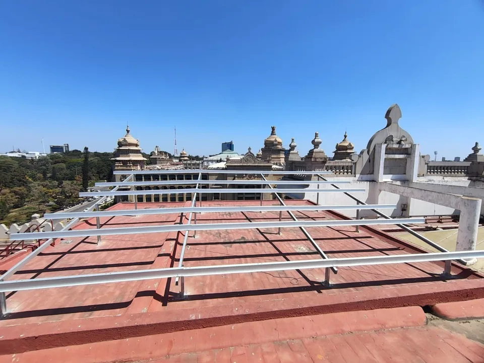 A low galvanised mounting structure fixed to a red-oxide terrace, with the domes and turrets of the Vidhana Soudha rising behind it