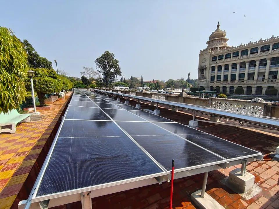 A long row of modules running beside a paved terrace walkway with benches and planting, the domed tower of the Vidhana Soudha behind
